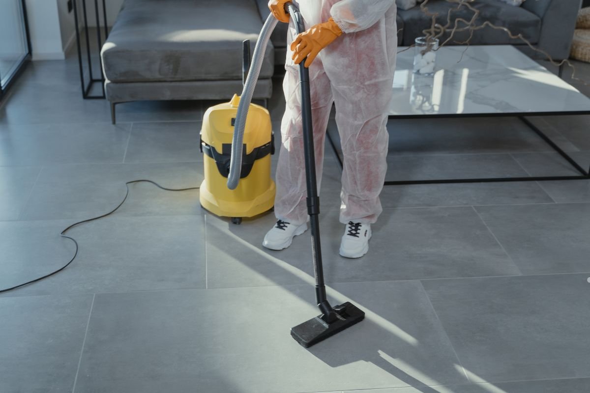 A person in protective gear vacuums a modern gray tile floor, using a yellow vacuum cleaner while standing near a couch.