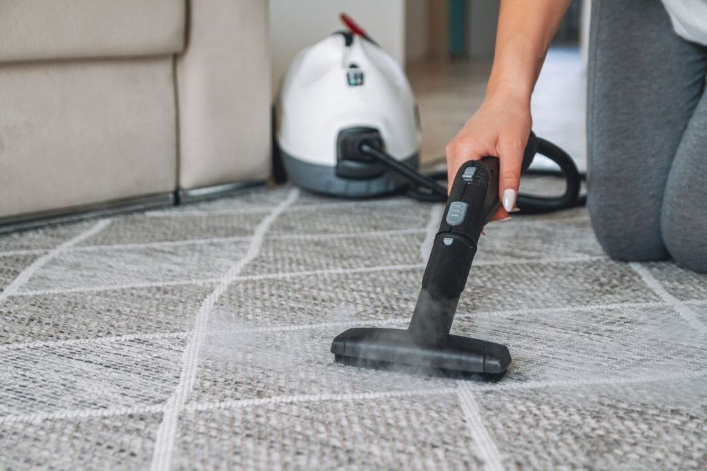 A person using a steam cleaner on a patterned carpet, with a white steam cloud, while a vacuum cleaner is visible in the background.